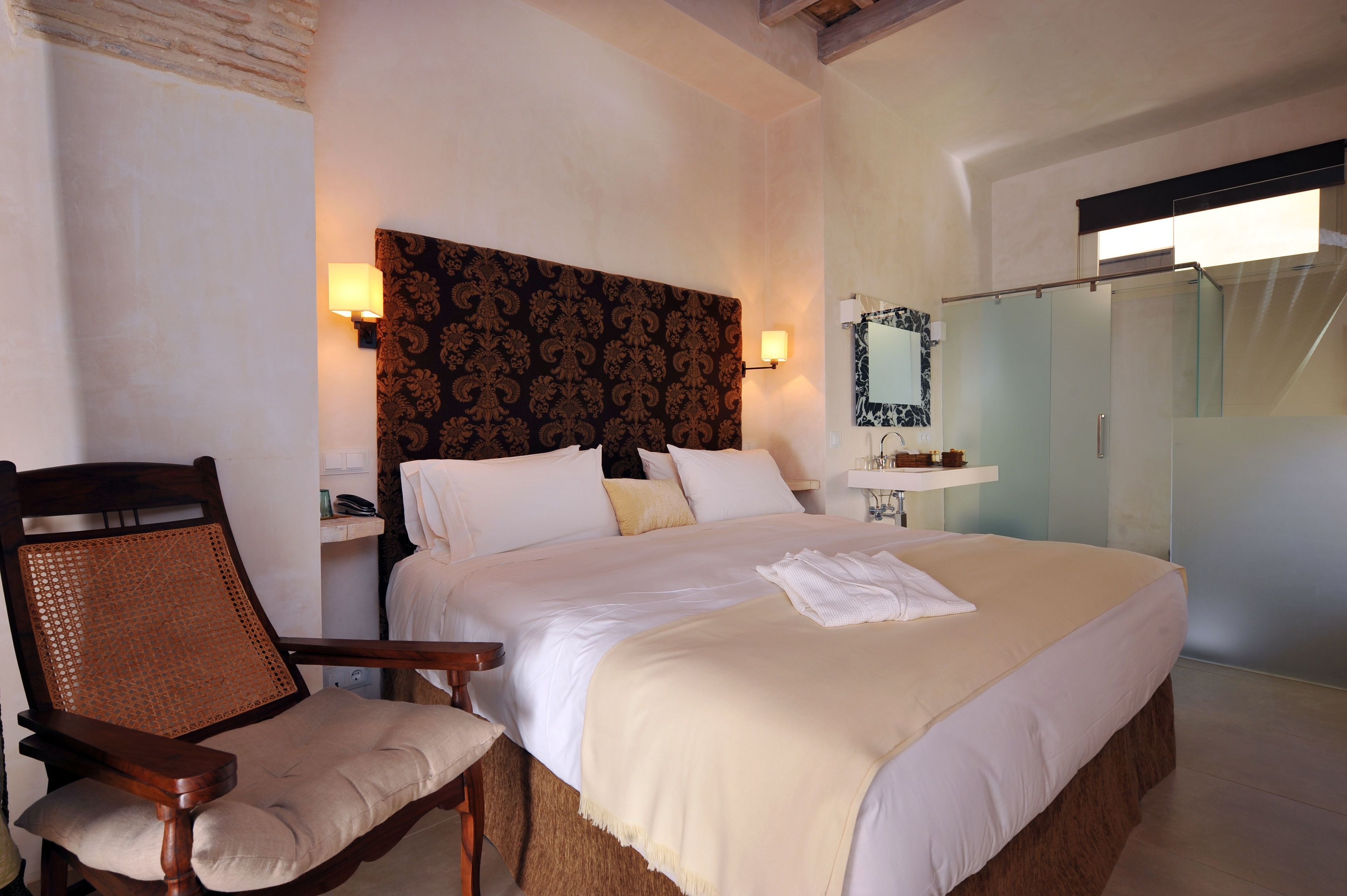 A hotel room with a large bed, a wooden chair, a nightstand with a lamp, and a bathroom with a glass shower.