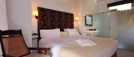A hotel room with a large bed, a wooden chair, a nightstand with a lamp, and a bathroom with a glass shower.