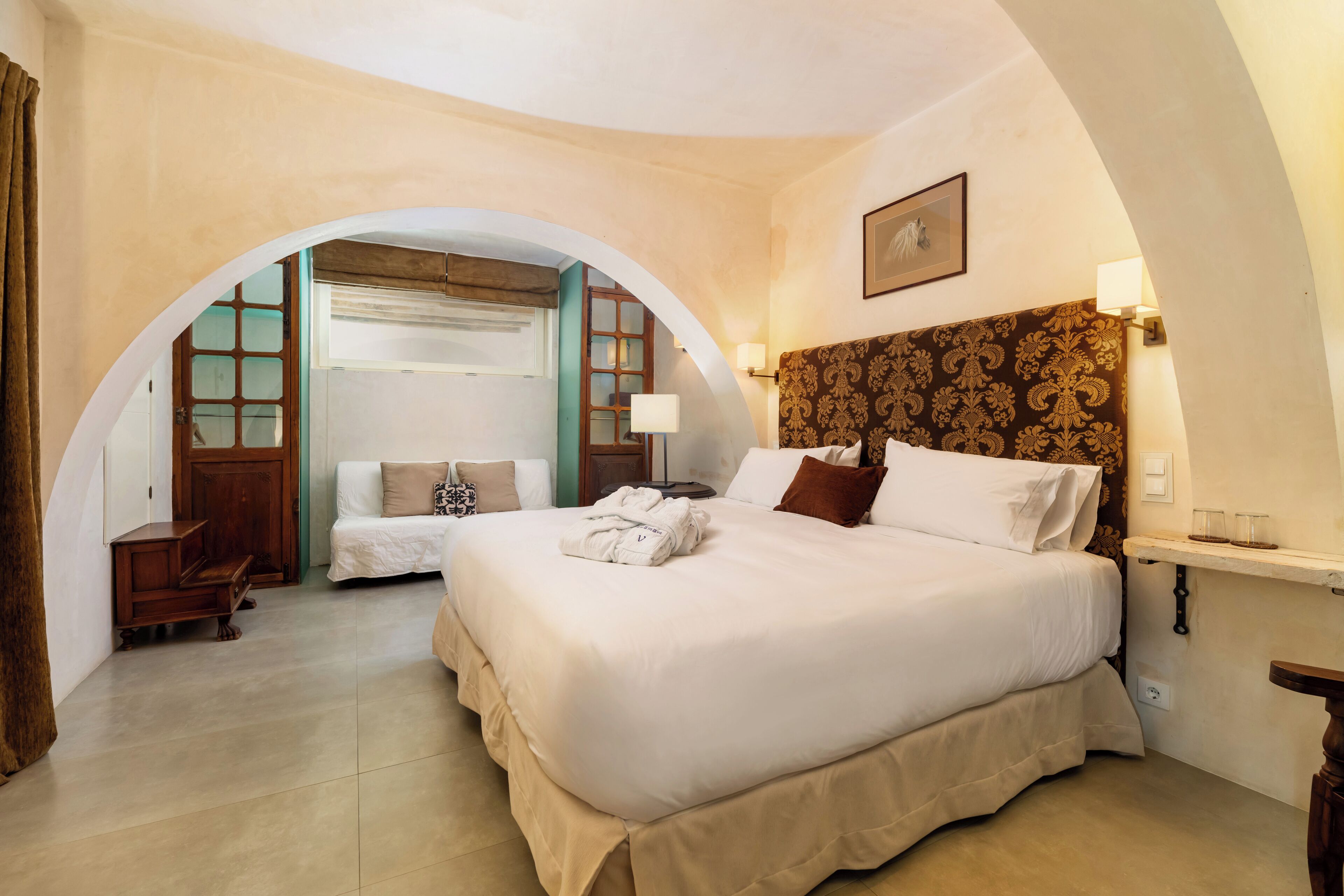 Une chambre d’hôtel avec un grand lit, un petit bureau, une chaise et une porte en arc donnant sur une autre pièce.