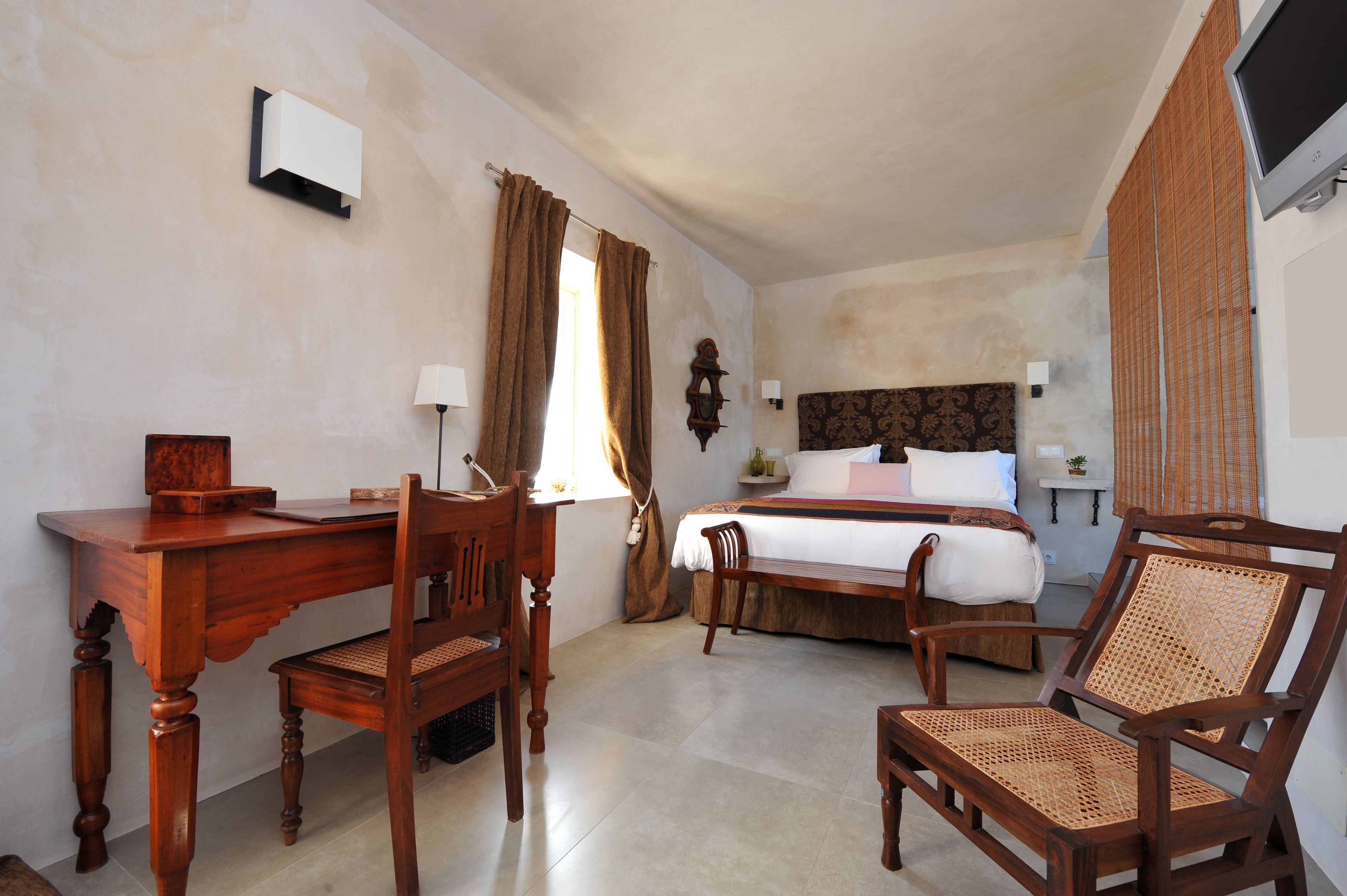 Habitación de hotel con escritorio de madera, dos sillas, cama con cubrecama blanco, mesita de noche con lámpara y televisor montado en la pared.