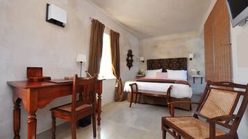 Habitación de hotel con escritorio de madera, dos sillas, cama con cubrecama blanco, mesita de noche con lámpara y televisor montado en la pared.
