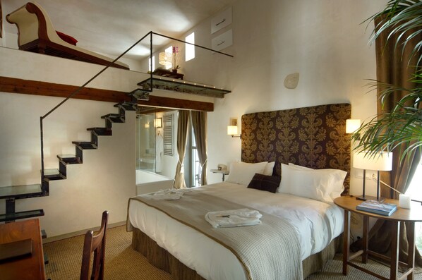 Hotelkamer met een bed, nachtkastje, lamp, stoel, bureau, spiegel, trap en een plant.