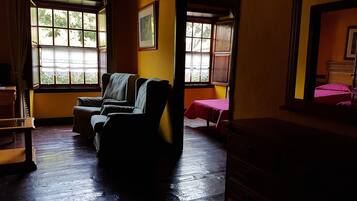 Una habitación con piso de madera, un sofá, una cama con colcha roja y un aparador. Hay ventanas con cortinas y un cuadro en la pared.