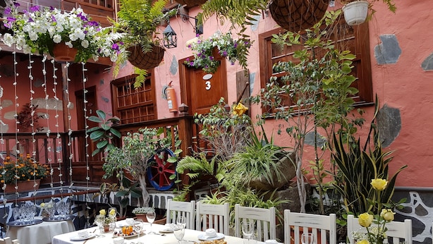 Un área de comedor al aire libre con mesas dispuestas para una comida, rodeada de plantas en macetas y decoraciones colgantes.