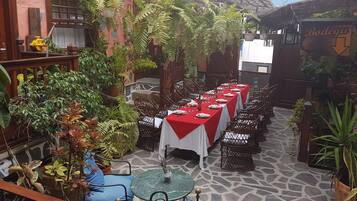 Un patio con una larga mesa de comedor lista para una comida, rodeado de plantas en macetas y muebles de mimbre.