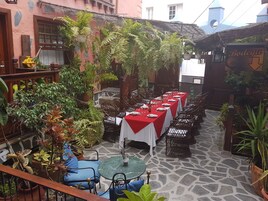 Un patio con una larga mesa de comedor lista para una comida, rodeado de plantas en macetas y muebles de mimbre.