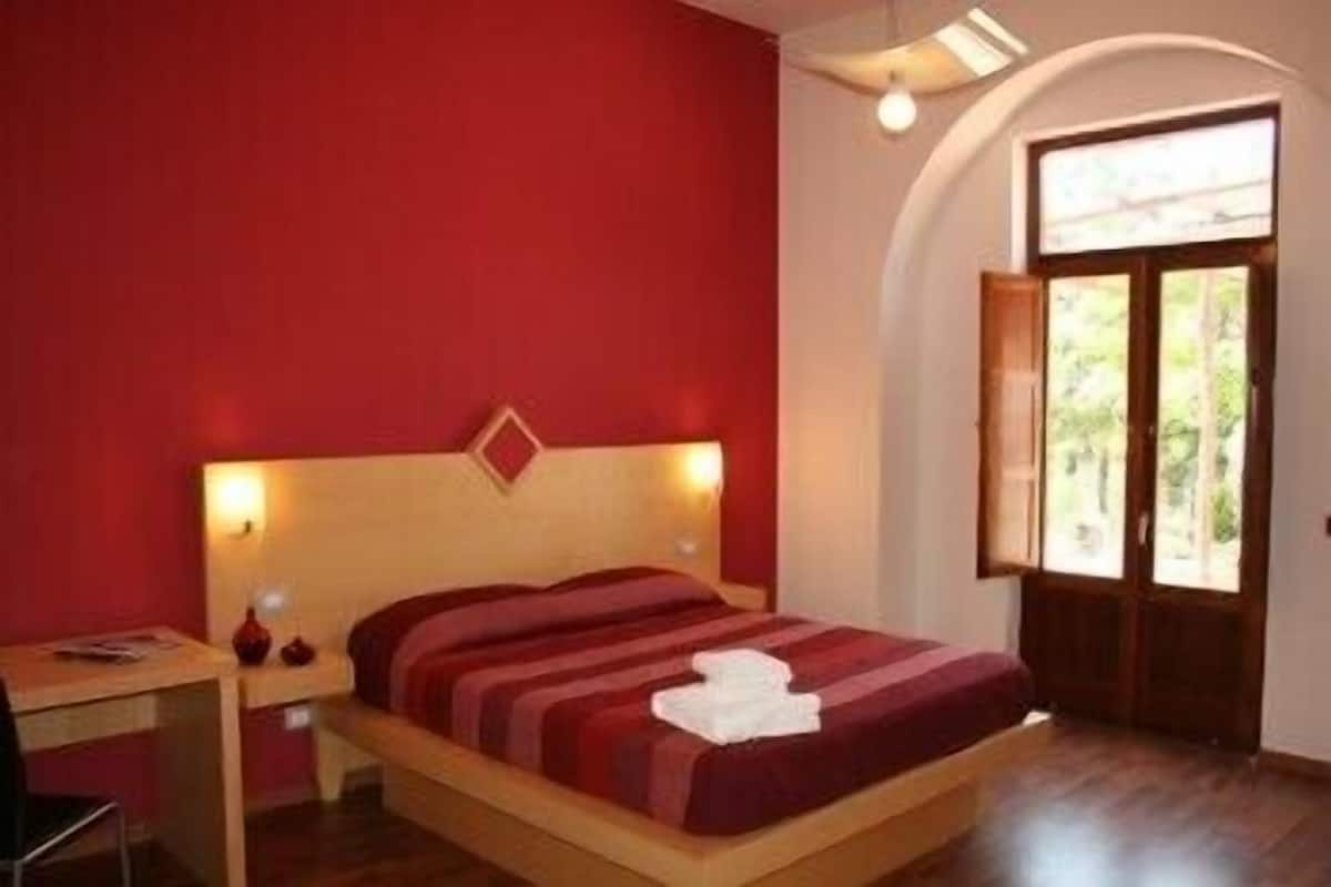 Une chambre à coucher avec un lit en bois, des murs rouges, un bureau avec une chaise et une fenêtre à arc.