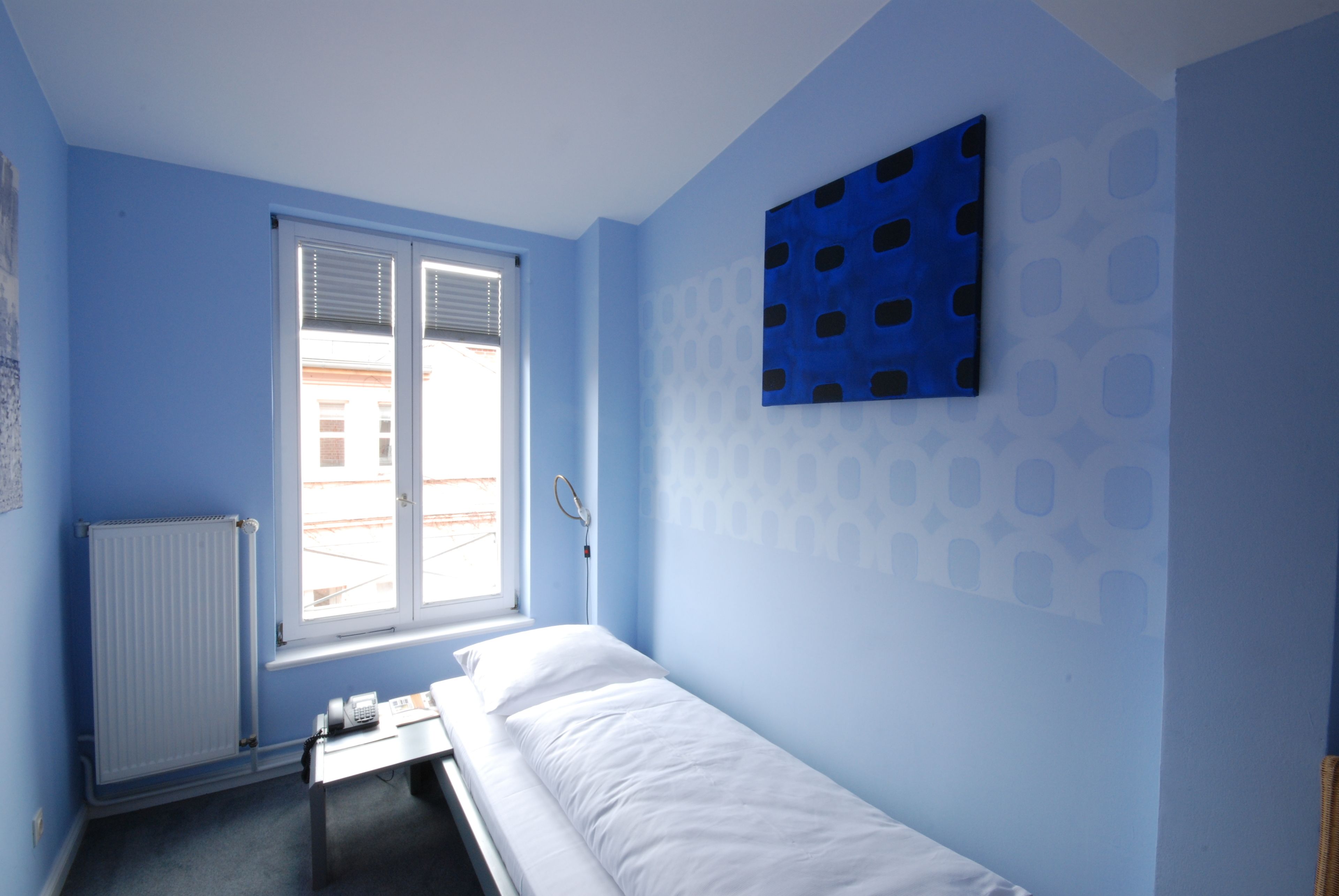 Una habitación pequeña con cama individual, ventana, radiador, mesita de noche y una obra de arte colgada en la pared.
