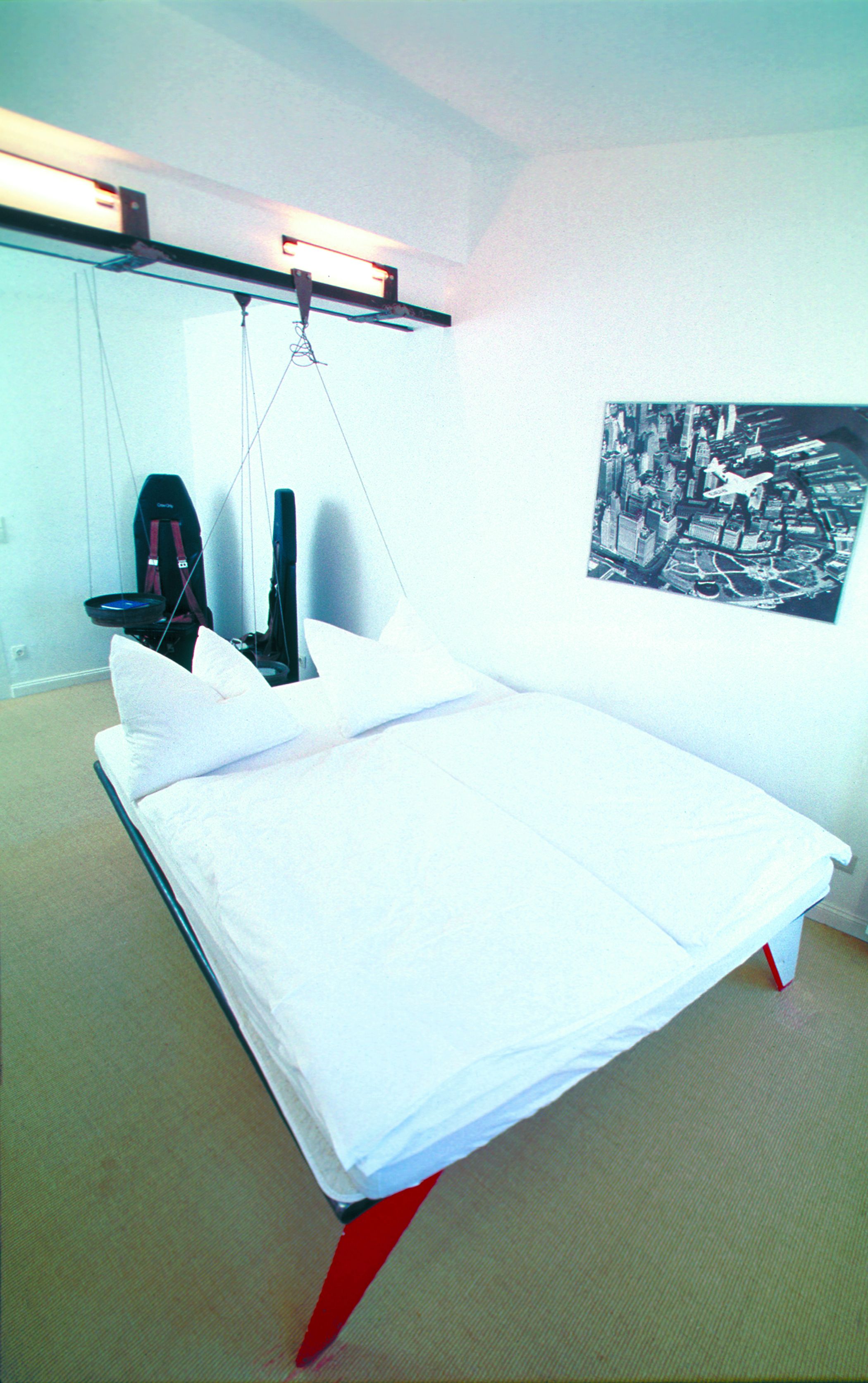 Una cama con ropa de cama blanca, un reposapiés rojo y una obra de arte blanco y negro en la pared.