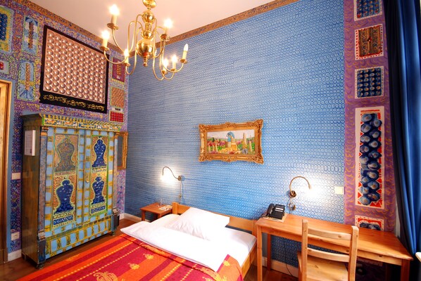 A room with a patterned blue wall, a bed with white linens, a wooden nightstand, a table with a lamp, and a chandelier. - Arte Luise – Self-Check In (Berlin)