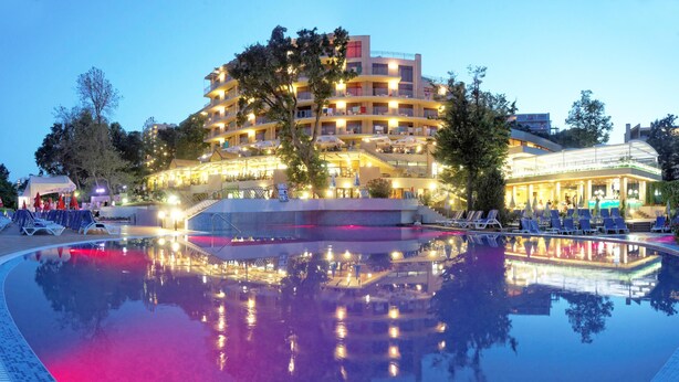 Et hotel med en pool, der spejler lysene, flere balkoner og udendørs siddeområder.