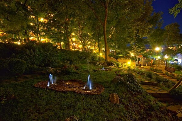 Un jardin nocturne doté de deux fontaines, entouré d’arbres et éclairé par un système d’éclairage d’ambiance.