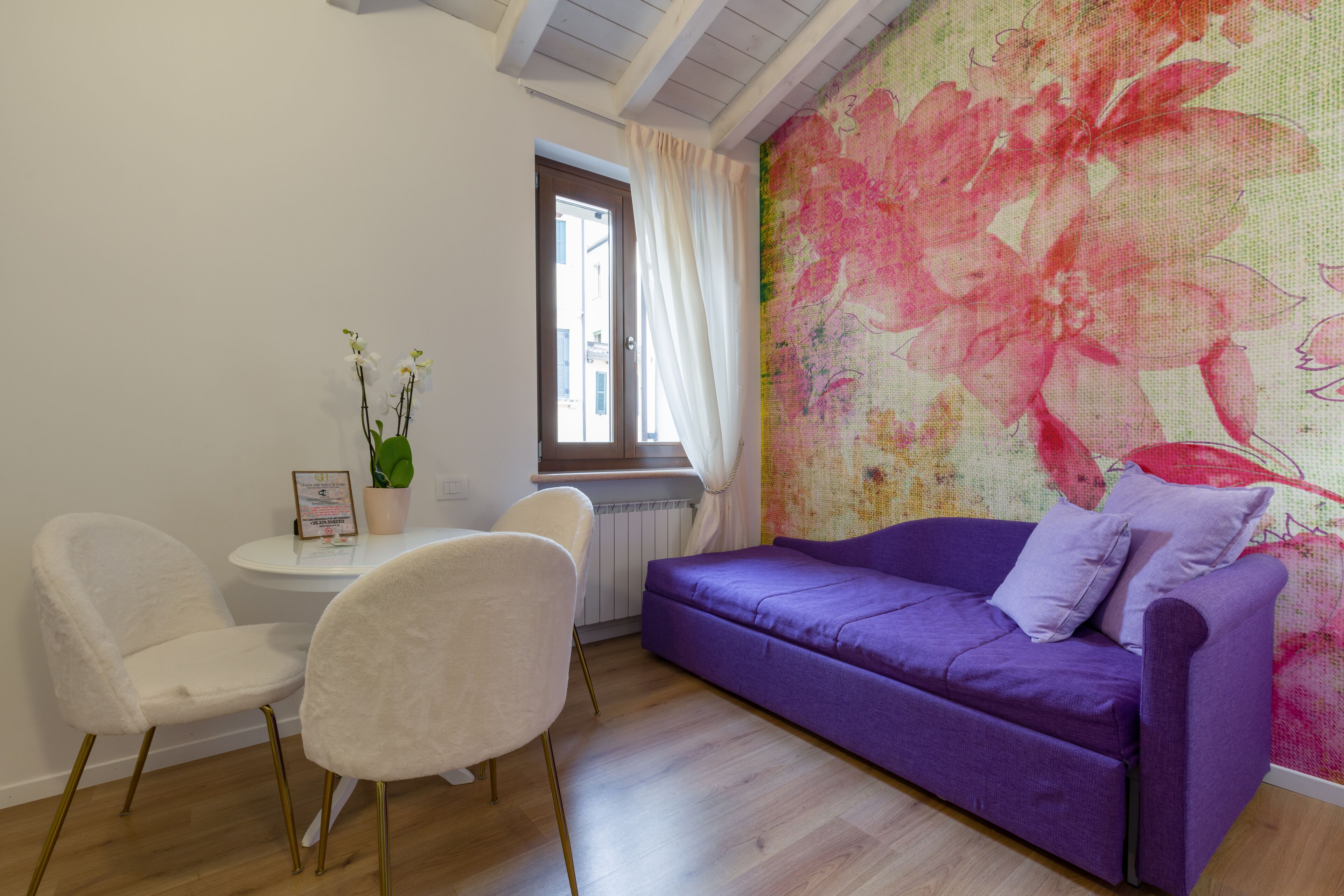 Une pièce avec un canapé violet, deux chaises blanches, une petite table avec une plante et une fenêtre avec des rideaux.