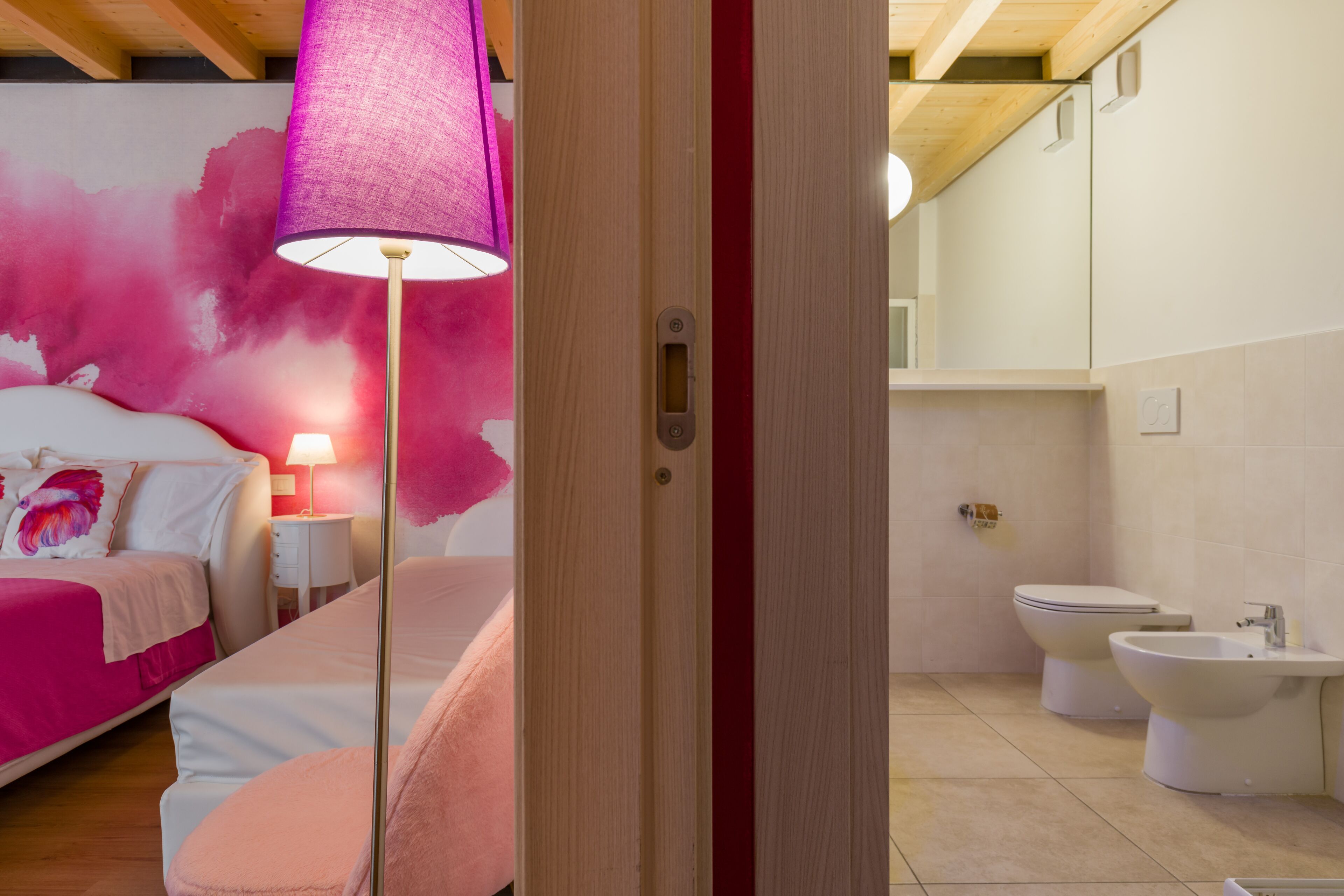 A bedroom with a bed, bedside table, and a lamp. The bathroom has a toilet, bidet, and sink.