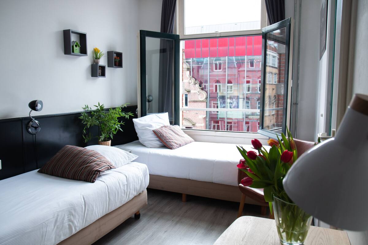 A modern hotel room with two beds, a large window, and a view of buildings outside.