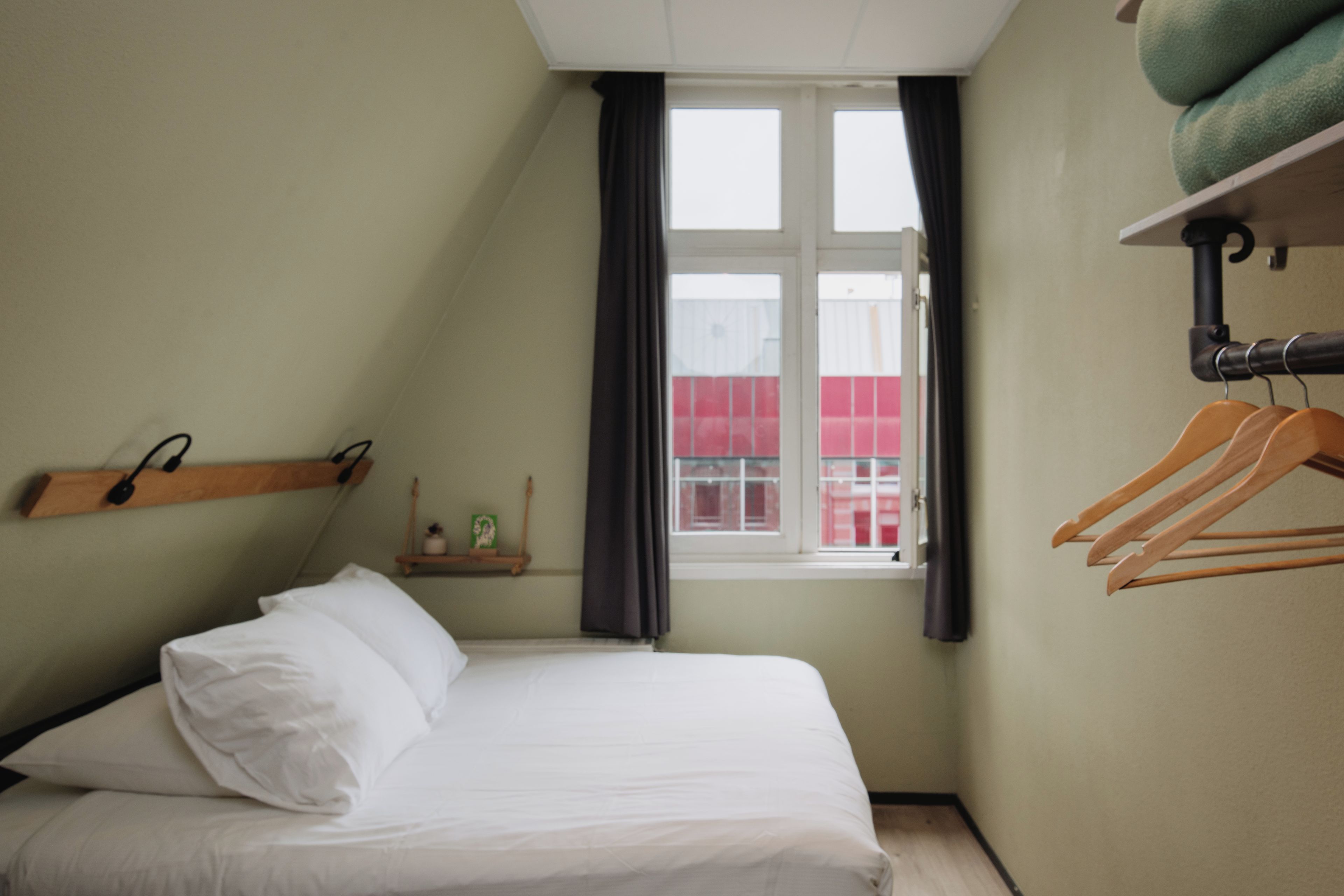 A bedroom with a bed, a window with curtains, a shelf with a plant, and a wall-mounted rack with hangers.
