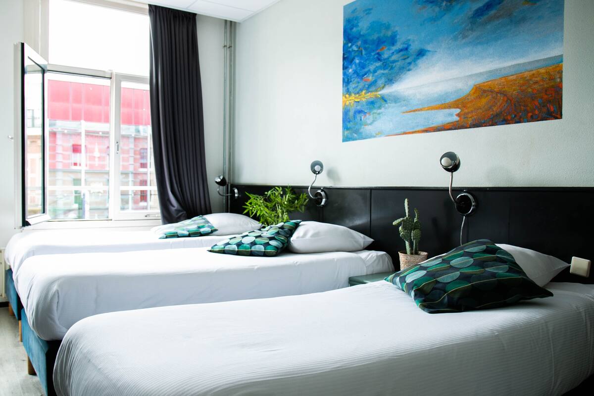 A hotel room with two beds, a large window with curtains, a painting on the wall, and a potted plant.