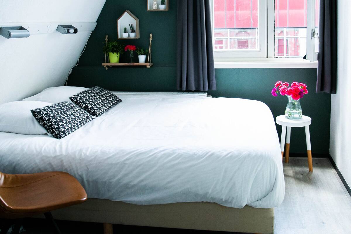 A bedroom with a bed, a chair, a window with curtains, a small table with a vase of flowers, and a shelf with plants and a mirror.