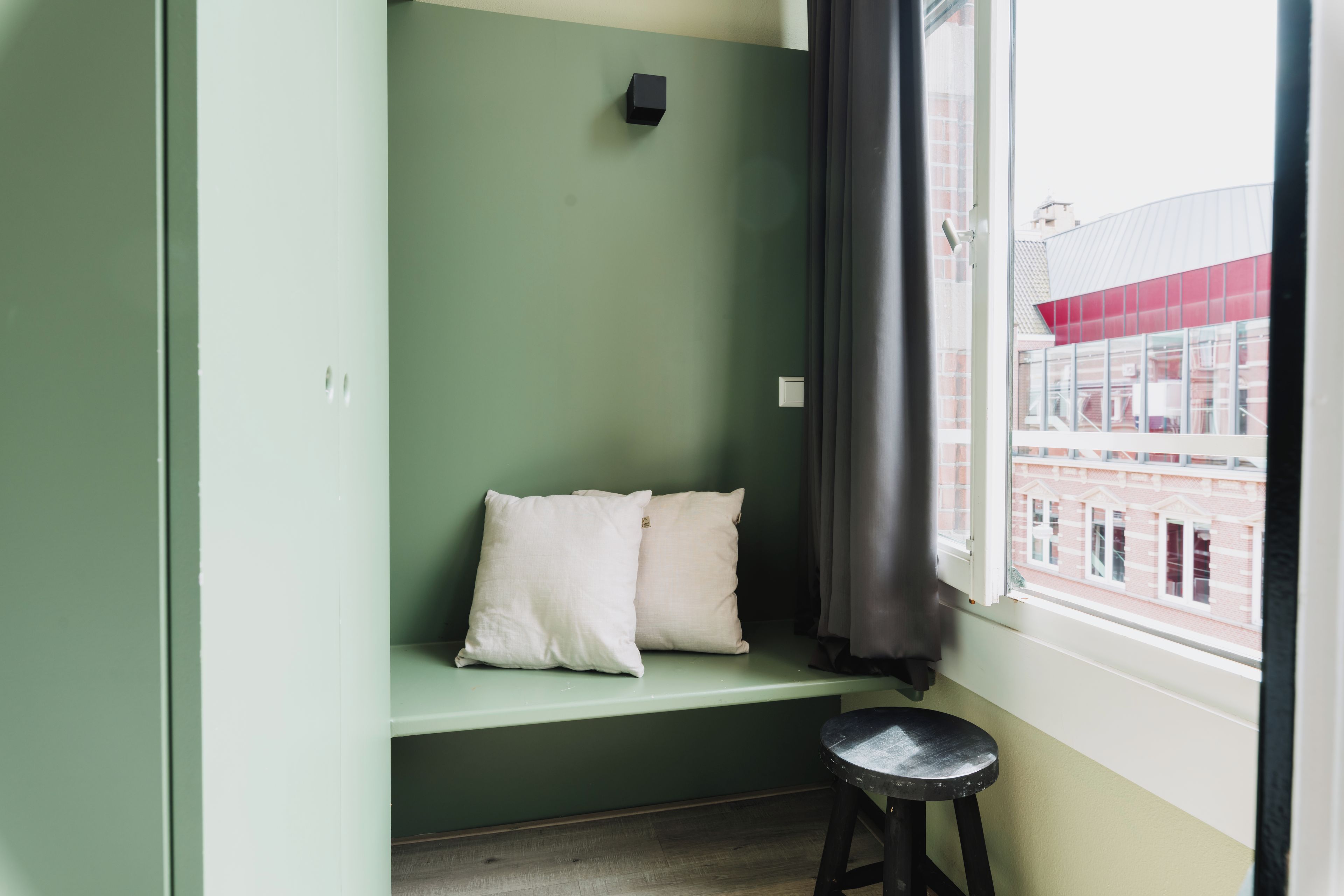 A room with a green wall, a window with a view of buildings, a black stool, and two pillows on a bench.