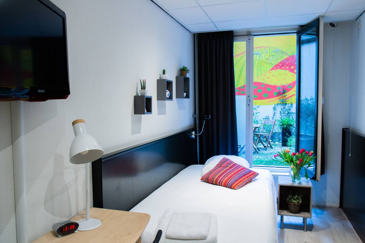 A hotel room with a bed, bedside table, lamp, TV, and a window with a view of a cityscape.