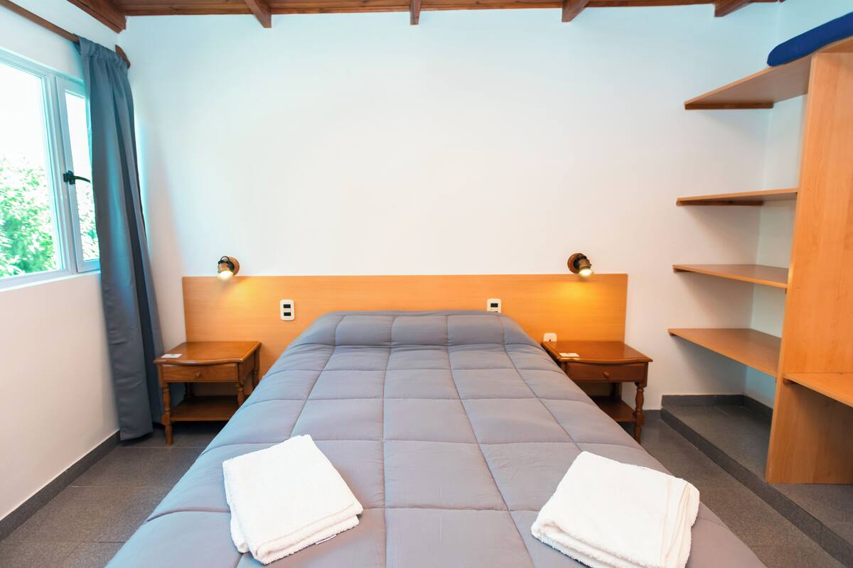 Une chambre comprenant un lit, deux tables de chevet, une commode, une fenêtre avec des rideaux et une étagère en bois.