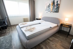 A hotel room with a bed, two bedside tables, a chair, a desk, and a cityscape painting on the wall.
