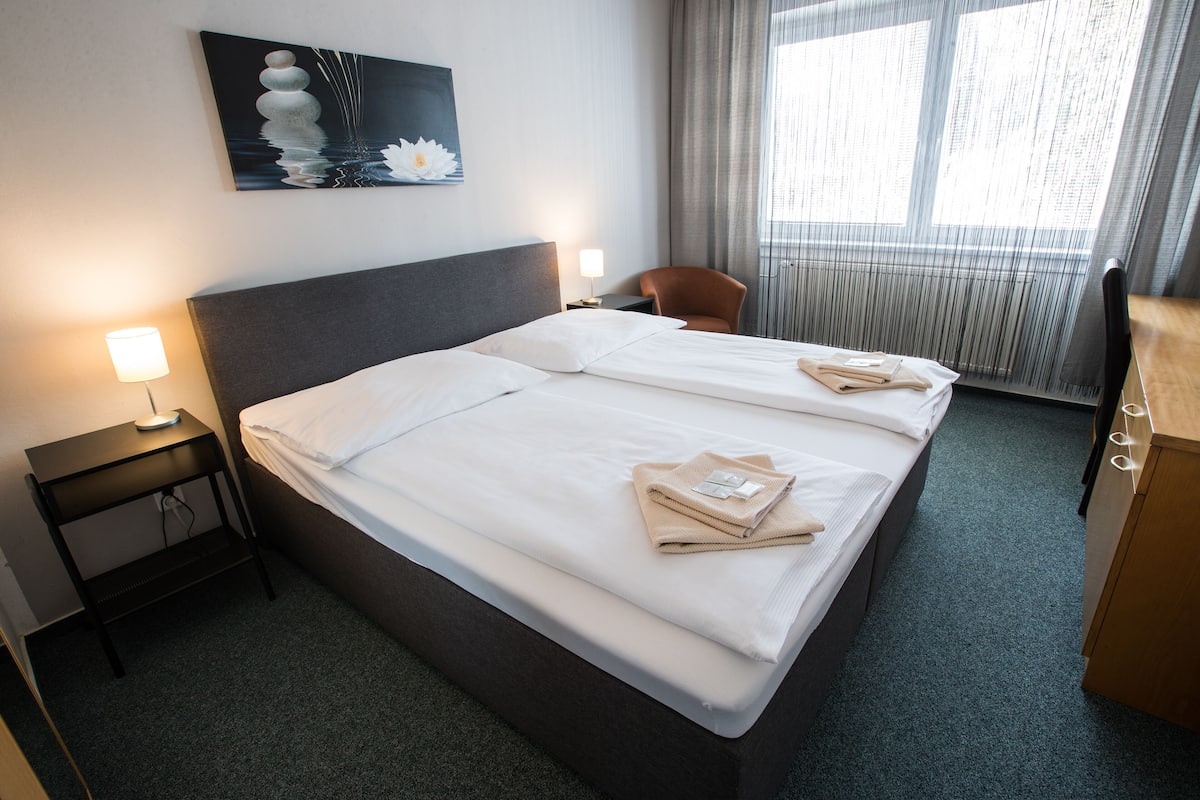 Une chambre d’hôtel avec un grand lit, deux lampes de chevet, une chaise, un bureau et une fenêtre avec des rideaux.