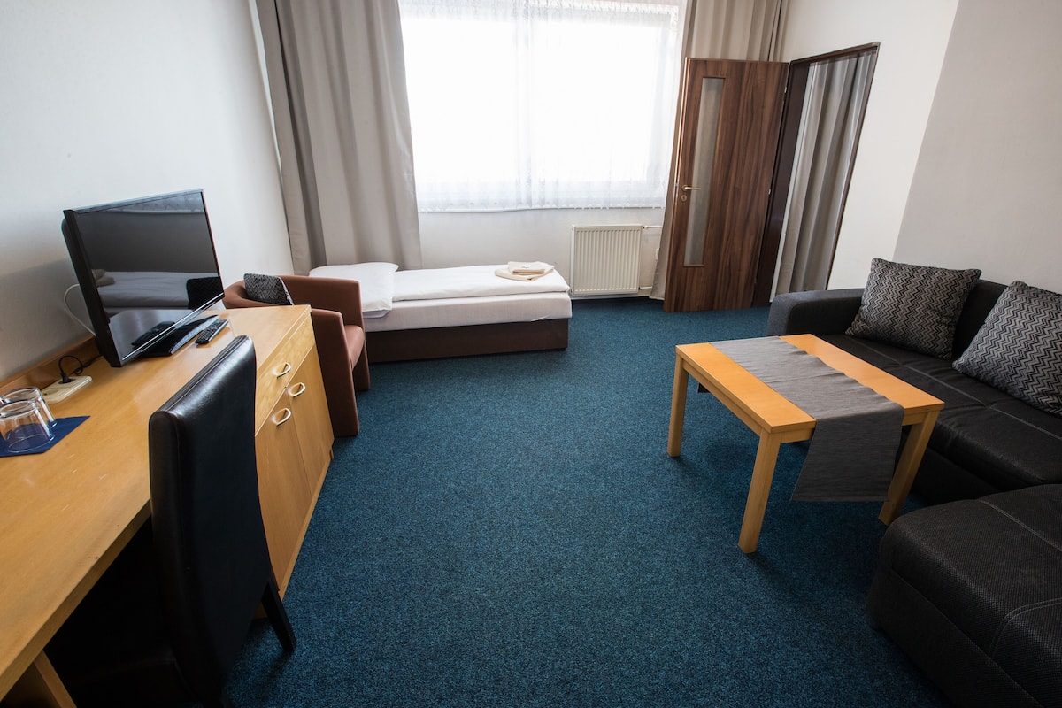 Une chambre d’hôtel avec un lit, un bureau avec un ordinateur, une petite table, un canapé et une fenêtre avec des rideaux.