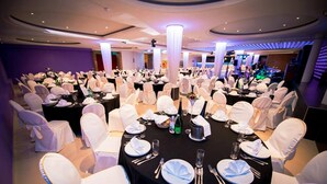 Une salle de banquet avec des tables rondes dressées pour un événement formel, agrémentée de nappes blanches, de chaises recouvertes de housses blanches et d’un design de plafond moderne.