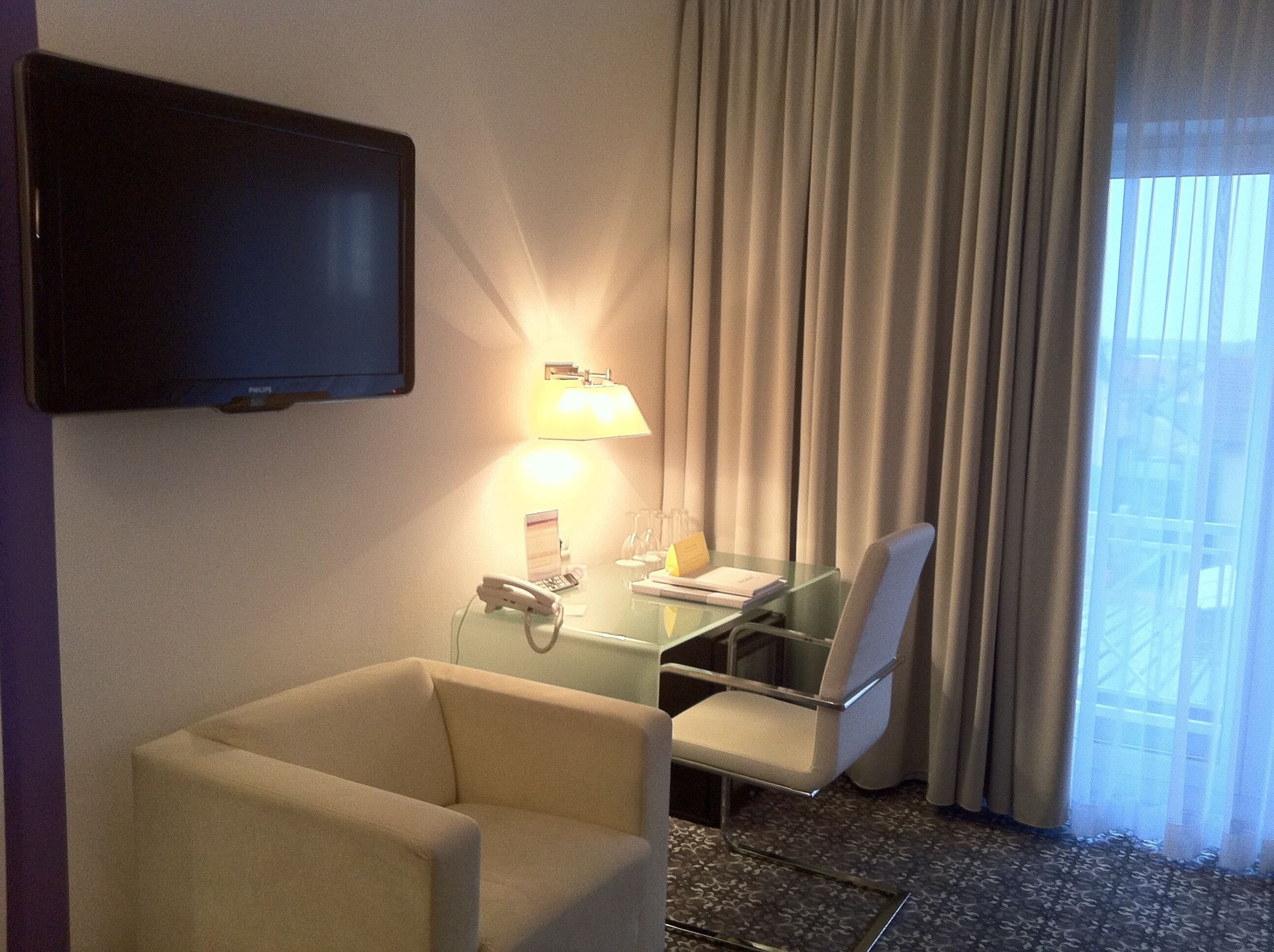 A hotel room with a flat-screen TV mounted on the wall, a glass desk with a chair, a lamp, and a telephone.