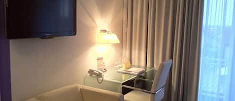 A hotel room with a flat-screen TV mounted on the wall, a glass desk with a chair, a lamp, and a telephone.