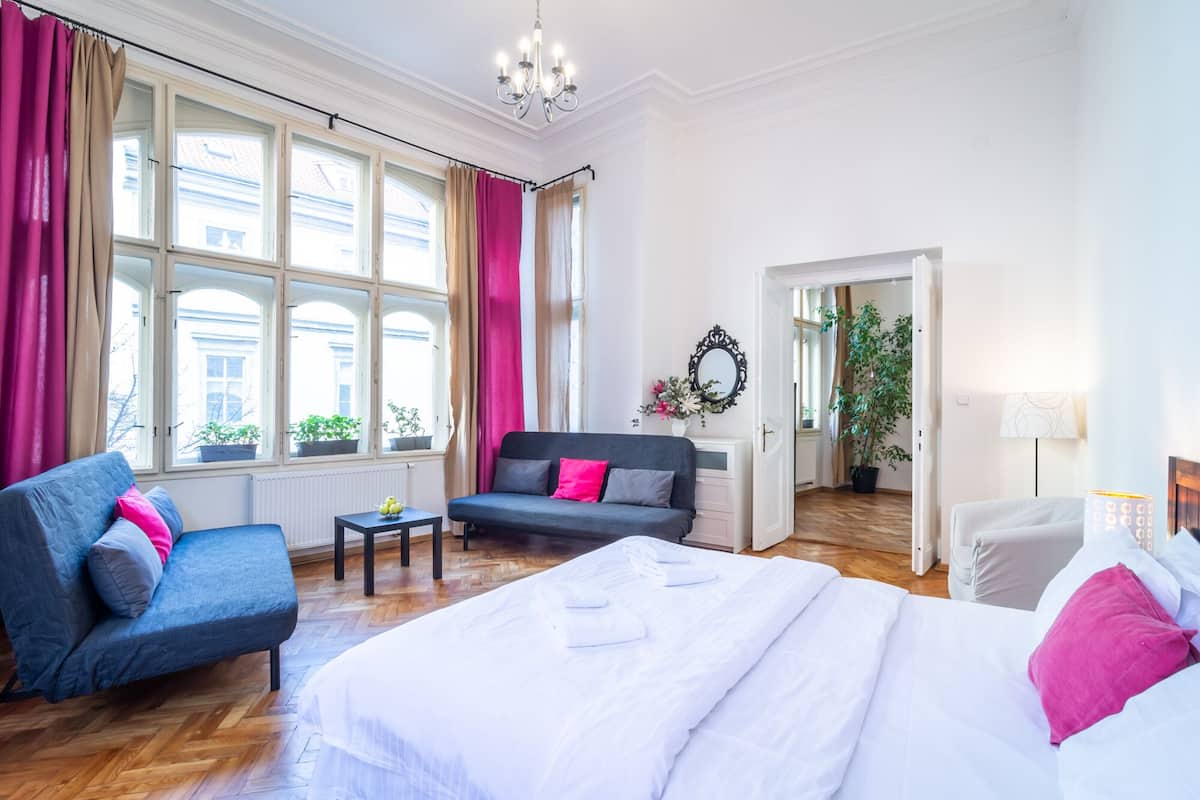 Camera d'albergo con un letto grande, una poltrona, un tavolino e una sedia. Sono presenti finestre con tende, un lampadario e una pianta.