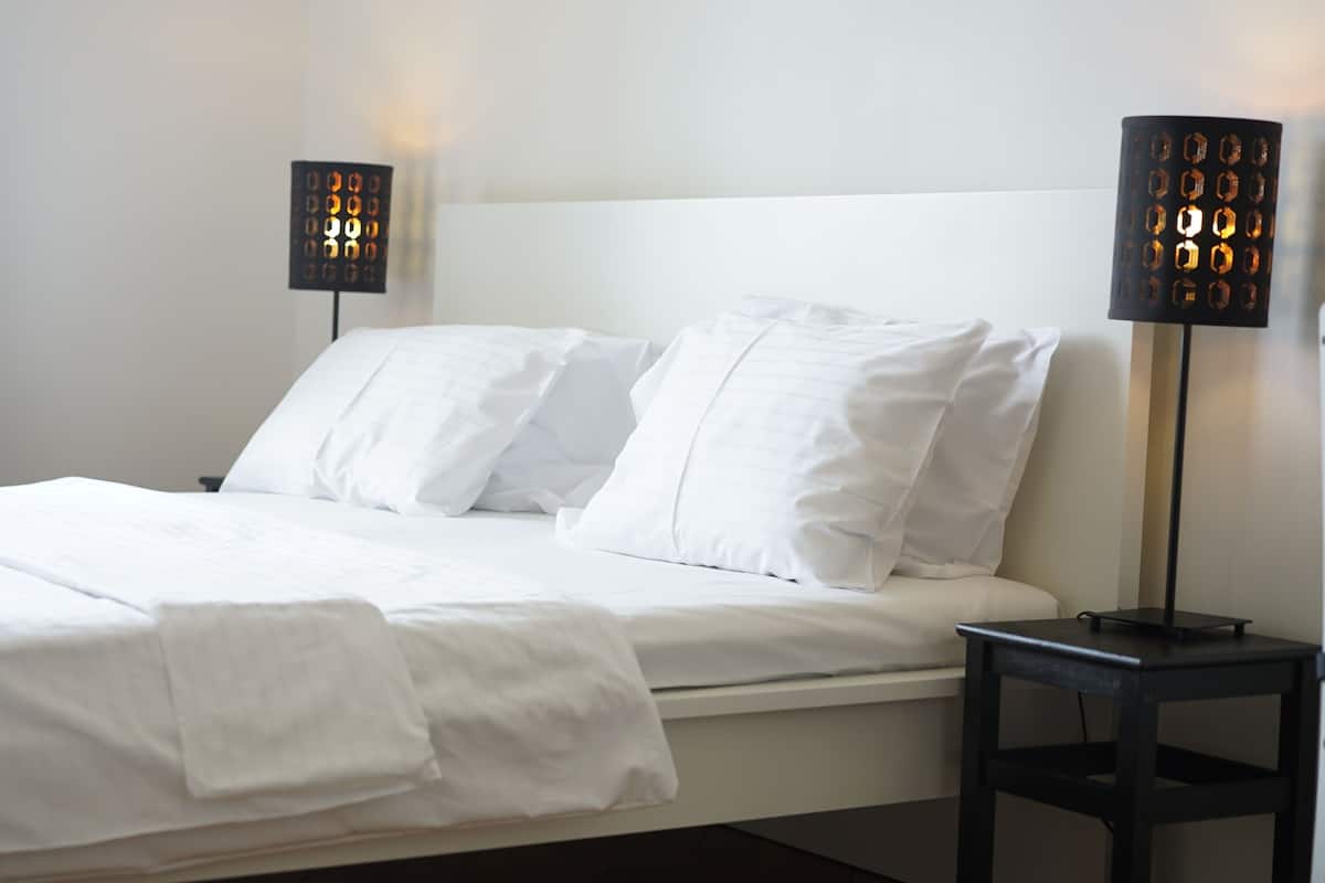 Un letto ben rifatto con lenzuola e cuscini bianchi, un comodino con una lampada e una finestra con tende.