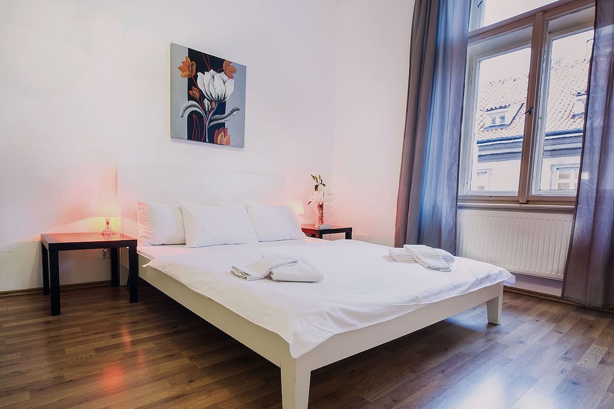 Una camera d'albergo con un letto grande, due comodini, un tavolino di notte, una finestra con tende e un quadro appeso al muro.