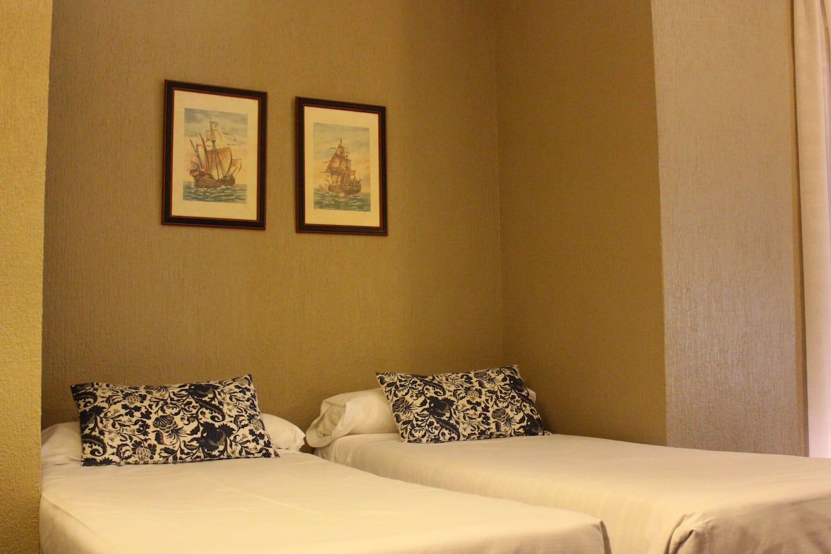 Dos camas individuales con almohadones estampados, una pared color beige y dos cuadros con imágenes de barcos.