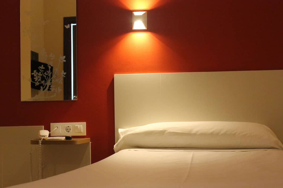 Habitación de hotel con cama, mesita de noche, lámpara de pared y toma de corriente.