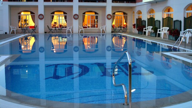 A swimming pool with a clear blue water surface, surrounded by white chairs and tables, and illuminated by warm interior lighting. - Hotel Caribe (Rota)