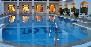 A swimming pool with a clear blue water surface, surrounded by white chairs and tables, and illuminated by warm interior lighting. - Hotel Caribe (Rota)