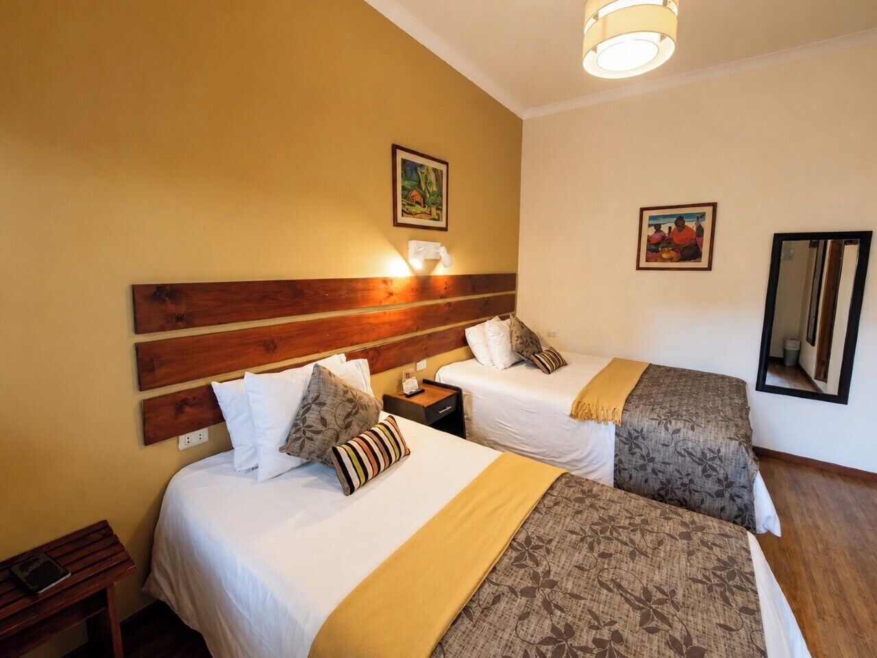A hotel room with two beds, a wooden headboard, wall-mounted lamps, and framed artwork.