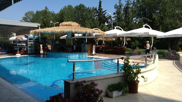 A swimming pool with a thatched-roof bar area, surrounded by white umbrellas and potted plants. - Flamingo (Nessebar)