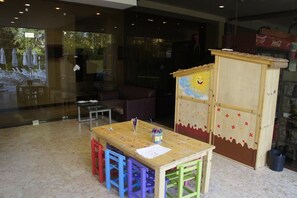 A children's play area with a wooden table and colorful chairs, a small playhouse, and a glass wall with outdoor seating visible.
