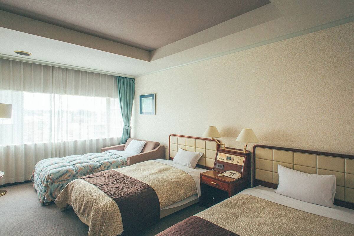 Una camera d'albergo con due letti, un comodino con lampade, una finestra con tende e un quadro appeso al muro.