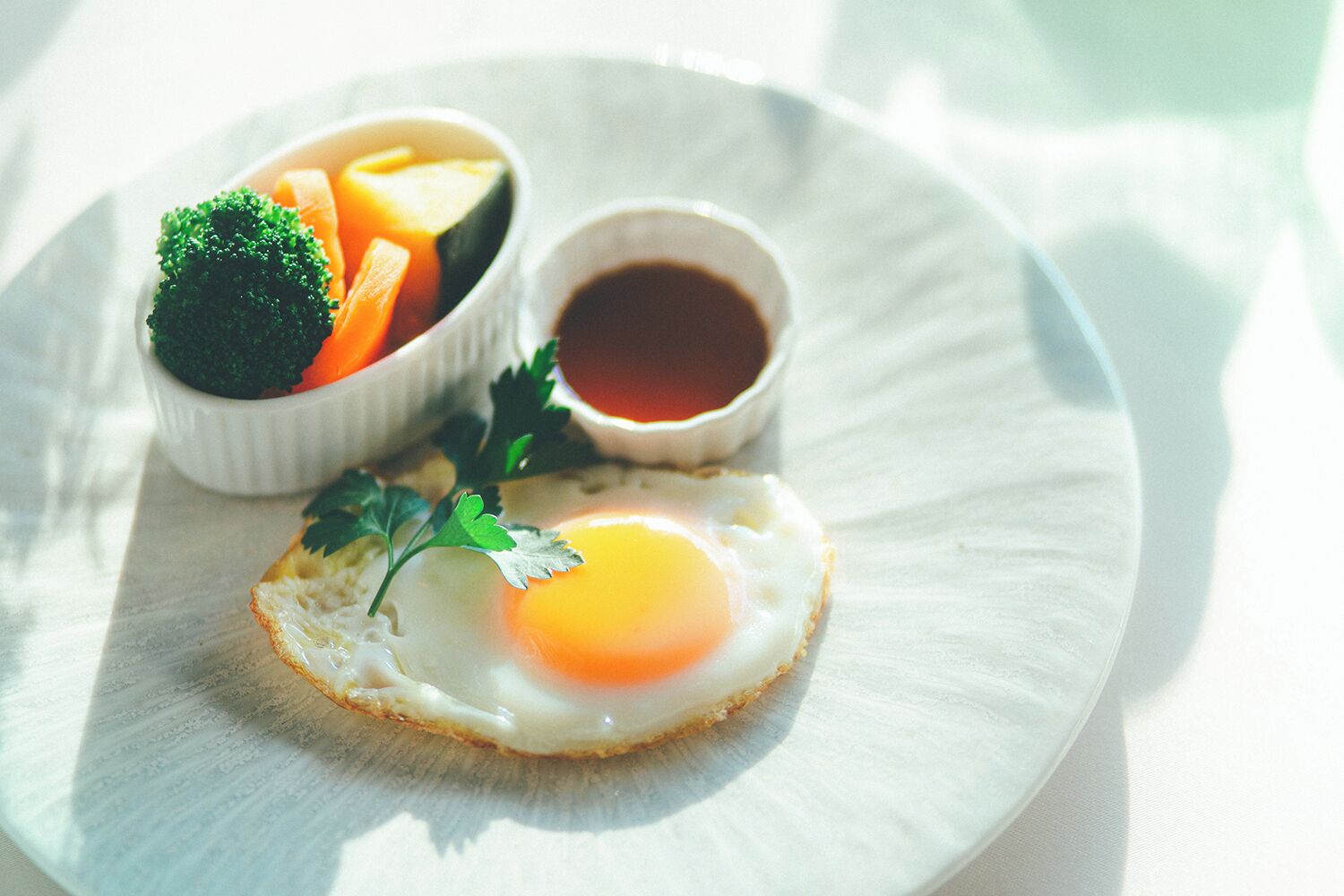 A plate with a sunny side-up egg, a bowl of mixed vegetables, and a small cup of sauce.