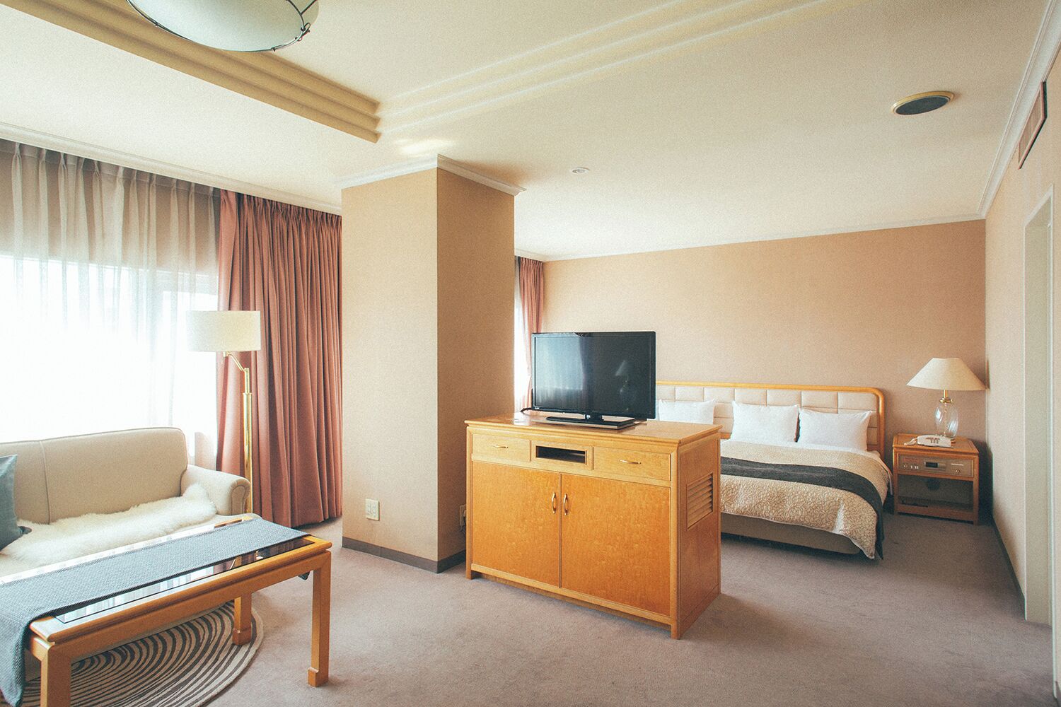 A hotel room with a bed, a TV on a wooden cabinet, a sofa, a lamp, and a bedside table.