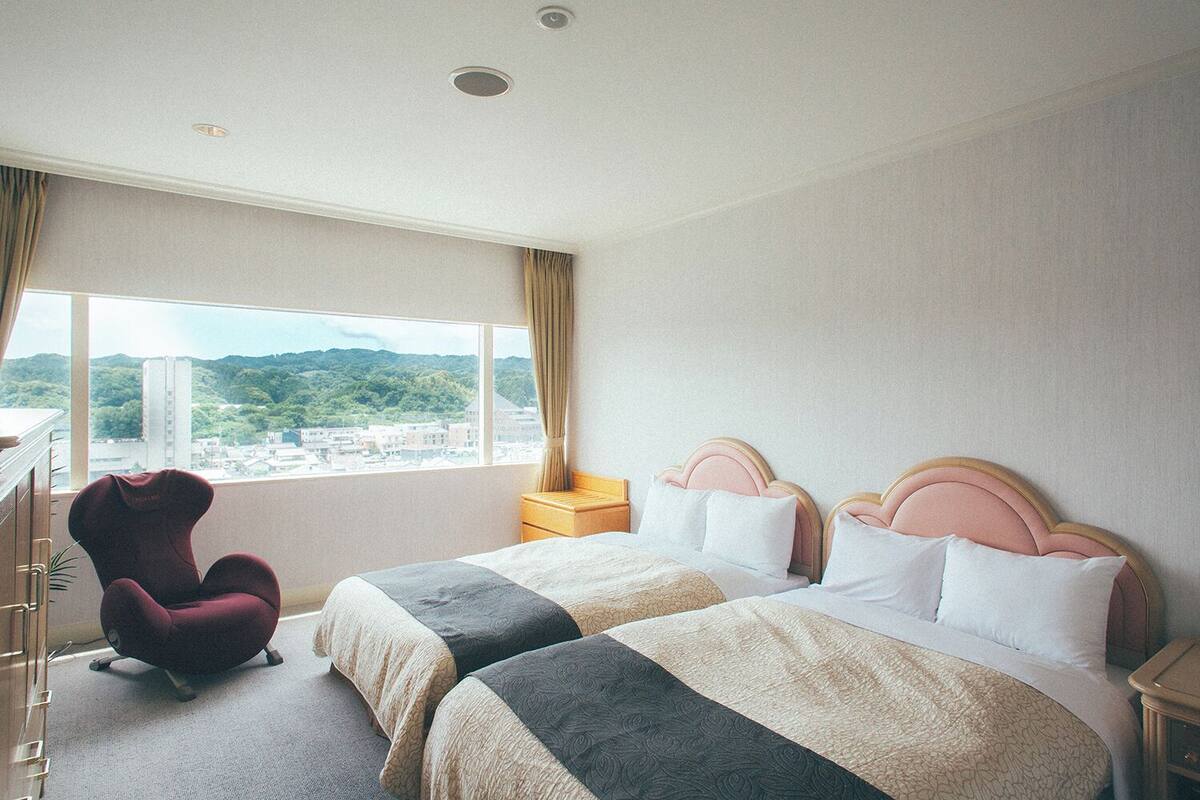 Una camera d'albergo con due letti, una sedia, una scrivania e un'ampia finestra con vista sulla città.