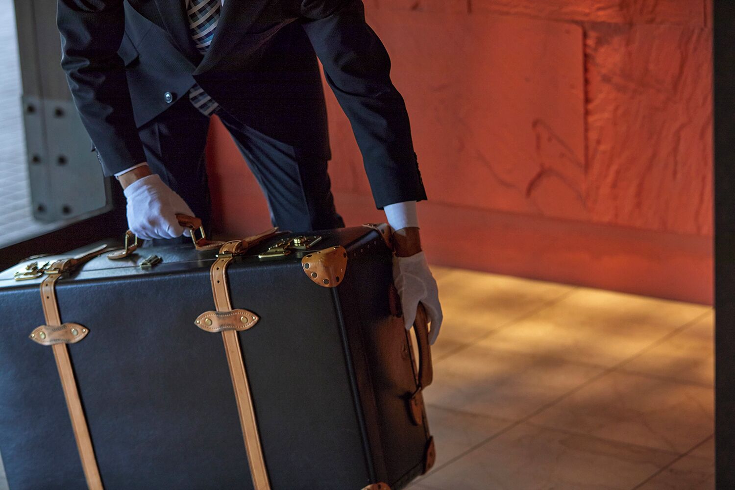 Une personne en costume, tenant une grande valise noire munie de sangles en cuir et de fermoirs métalliques.