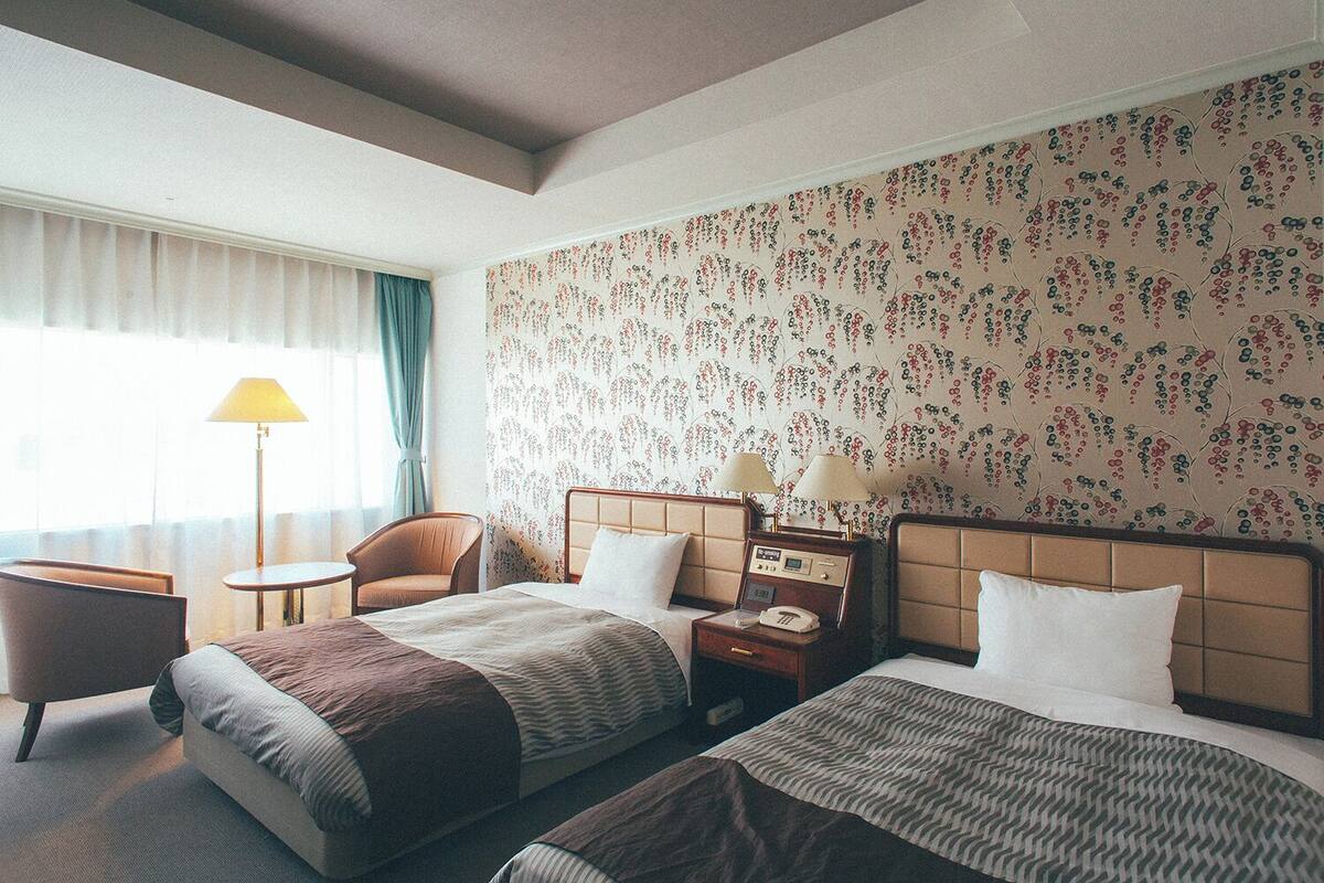 Una camera d'albergo con due letti, una sedia, un tavolino e una lampada.