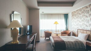 Quarto de hotel com uma cama, uma escrivaninha com abajur, televisão, duas cadeiras, uma mesinha e uma janela com cortinas.
