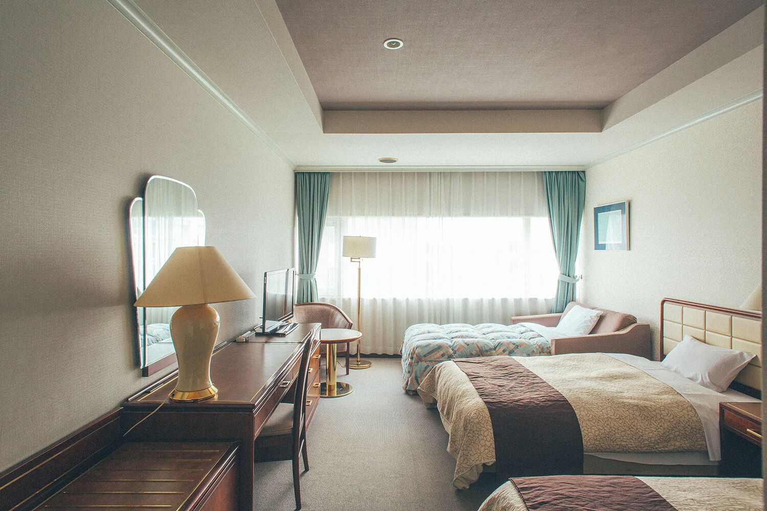 Habitación de hotel con escritorio, dos camas, una silla, lámpara y un ventanal con cortinas.