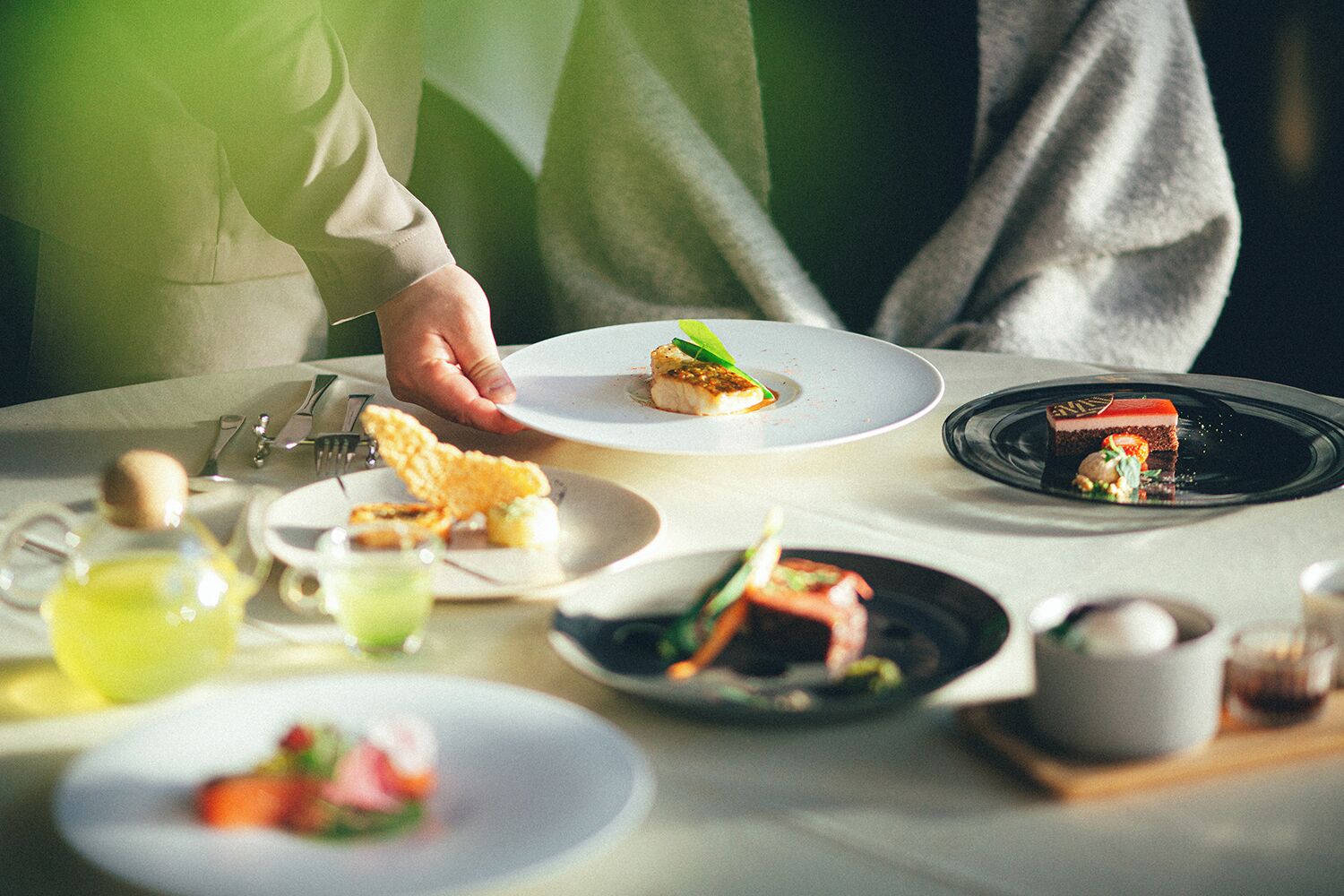 A dining table with various dishes, including a dessert on a black plate, a plate with a fish dish, and a plate with a green vegetable dish.